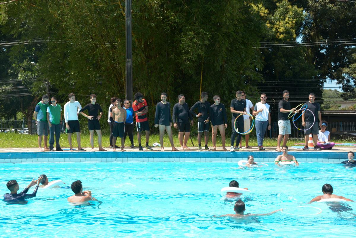 estudantes dentro da piscina, com ajuda de boias, e outros estudantes fora dela atirando bambolês para acertar nos que estao na piscina