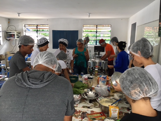 Participantes na cozinha, fazendo receitas, todos com toucas, ao redor de uma mesa com ingredientes