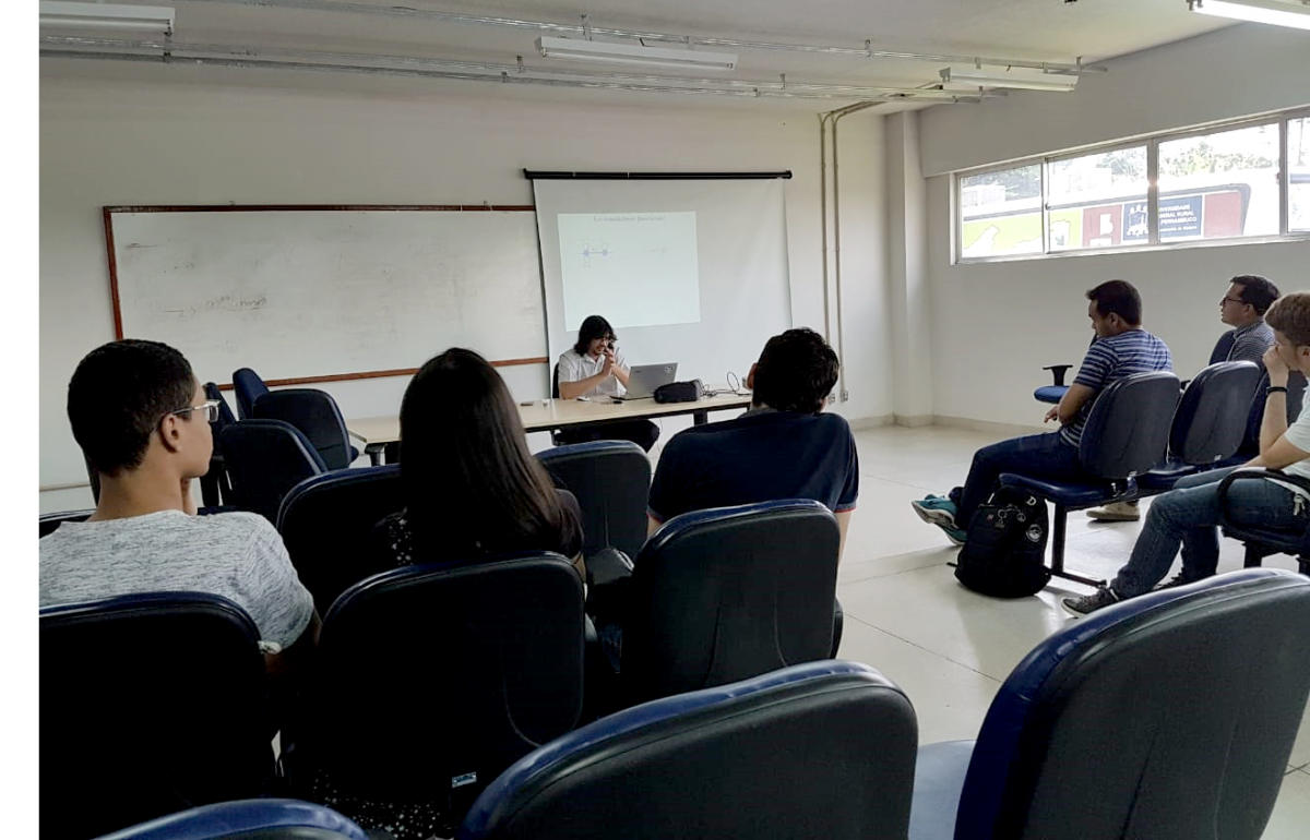 professor ministrando palestra sentado em birô. Estudantes assistem em suas respectivas cadeiras